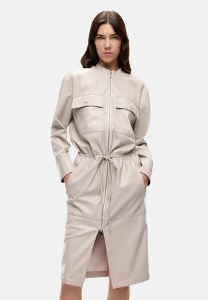 Mujer que lleva un vestido de piel sintética color beige claro con cremallera, mangas largas, bolsillos en el pecho, cintura elástica con cordón y abertura frontal.