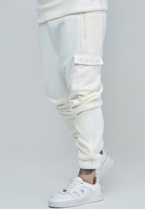 Hvide skrånende cargojoggere med sidelomme og lynlås, båret med hvide sneakers og med en tatoveret ankel, der vises.