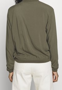 Olive green lightweight jacket with elastic cuffs and hem. Soft fabric, minimal design. Worn with white pants, showing back view.
