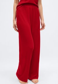 Γυναίκα που φοράει χαλαρό, κόκκινο πλεκτό παντελόνι με ριγωτή υφή και ταιριαστό τοπ, όρθια ξυπόλυτη μπροστά σε απλό ανοιχτό φόντο.