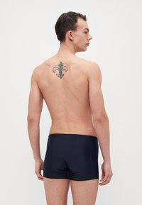 Marinefarbene Schwimmshorts mit passgenauer Passform, glatter Textur und elastischem Bund, getragen von einem Modell mit sichtbarem Tattoo auf dem Rücken.