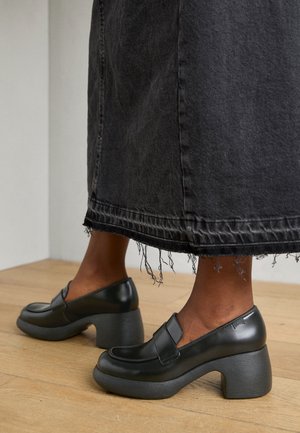 Mustad nahast block-kontsad kingad ümarate varvaste ja õmblusdetailidega. Kandmise alla on valitud narmastega äärega tume denim seelik puidust põrandal.