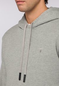 Šedá bavlněná mikina s velkou klokaní kapsou, stahovacími šňůrkami a malým vyšívaným logem na hrudi. Měkká textura s uvolněným střihem.