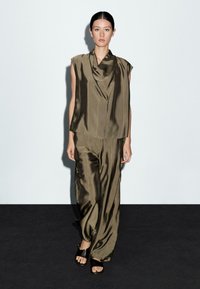 Vrouw loopt over een zwarte catwalk in een olijfgroen satijnen mouwloos top met gedrapeerde halslijn en bijpassende wijde broek, met zwarte sandalen.