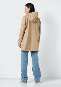 Manteau imperméable beige avec capuche, doté d'un design élégant et de finitions sur les coutures latérales, associé à un jean bleu clair à jambes larges.