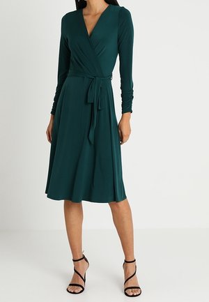 Femme portant une robe portefeuille vert foncé longueur genou avec des manches longues froncées et des sandales noires à talons hauts à brides, debout sur un sol blanc.