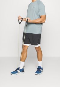 Blå sportskor, grå t-shirt och svarta shorts. Personen håller i träningsgummiband med svarta handtag och står framför en vit bakgrund.