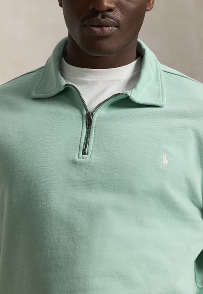 Άνδρας φορώντας ένα φούτερ με φερμουάρ μέχρι το στήθος σε πράσινο μέντας πάνω από ένα λευκό μπλουζάκι με στρογγυλή λαιμόκοψη, εμφανίζοντας το φερμουάρ και το κεντημένο λογότυπο στο στήθος.