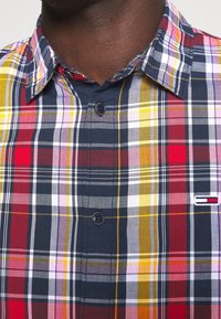 Uomo che indossa una camicia a quadri colorata con colletto, caratterizzata da strisce rosse, gialle, blu navy, bianche e viola, e un piccolo logo rettangolare sul petto.