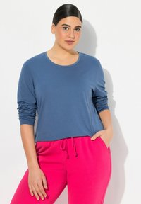 Modrá košile s dlouhým rukávem a kulatým výstřihem, zkombinovaná s růžovými kalhotami na stažení. Materiál vypadá měkce, s mírně nařasenými rukávy.