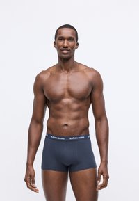 Καλά γυμνασμένος άνδρας που στέκεται μπροστά, φορώντας ναυτικές μποξερίτσες με λογότυπο Björn Borg στον ζωνάκι, σε απλή φόντο.