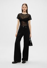 Top preto de malha transparente com riscas horizontais, combinado com calças pretas de cintura alta e perna larga. Os acessórios incluem uma pequena mala preta.