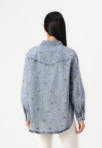 Ανοιχτό μπλε τζιν πουκάμισο με λουλουδάτη κεντητική, κουμπωτό μπροστινό μέρος, μακριά μανίκια και καμπυλωτό στρίφωμα. Διαθέτει κλασικό γιακά και μανίκια με γυριστές μανσέτες.