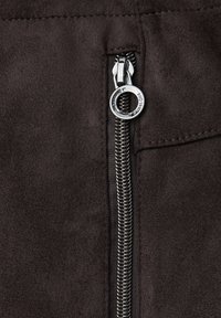 Brązowa zamszowa tkanina z detalami w kolorze srebrnym, z okrągłym uchwytem i szytymi krawędziami. Teksturowana powierzchnia i zbliżenie na detale.