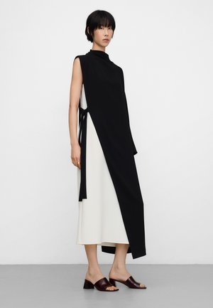 Vrouw staand in een zwart mouwloos asymmetrisch overlaagjurk die aan de zijkant is gestrikt, met daaronder een witte midi-jurk, en donkerkleurige mules met hakken.