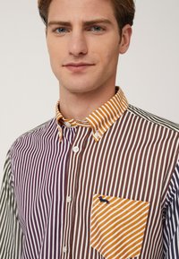 Camicia a righe con pannelli multicolore in viola, marrone e verde. Presenta una tasca a toppa con righe arancioni e bianche.