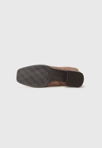 Talpa de pantof din suede maro, cu o talpă din cauciuc texturată, având modele în wave și un toc ușor. Logo-ul este vizibil pe latura tălpii.