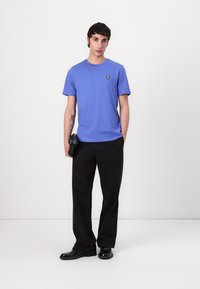 T-shirt bleu à manches courtes avec un petit logo jaune sur la poitrine, associé à un pantalon noir et des chaussures noires. Design et coupe minimalistes.