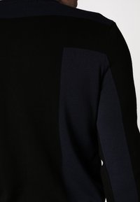 Pull en maille noir et marine avec une texture côtelée, présentant des blocs contrastants sur les épaules, offrant une silhouette ajustée.