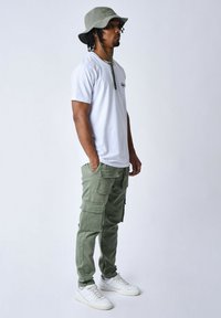 Camiseta blanca, pantalones cargo verde oliva con bolsillos y un sombrero tipo cubo a juego. Zapatillas blancas con suelas texturizadas completan el atuendo.