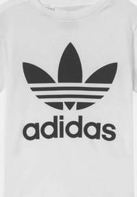Bijela pamučna majica s velikim crnim Adidas logotipom, tri pruge i grafika nalik listu iznad naziva branda.