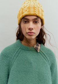 Turkoosi neulottu neulepaita, jossa on tekstuuroitu, väljähkö neulos; kerrostettu ruudullisella, kauluksellisella paidalla ja päällä keltainen kaapelineulottu pipo.