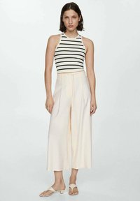 Biely a čierny pruhovaný crop top s vysokým výstrihom, kombinovaný s voľnými svetlo béžovými nohavicami. Outfit dokončujú jednoduché sandále.