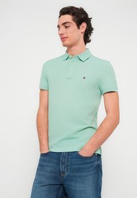 Ανοιχτό πράσινο πόλο πουκάμισο με γιακά, διαθέτει τρία κουμπιά και ένα μικρό λογότυπο στο αριστερό στήθος. Φοριέται με μπλε τζιν.