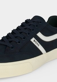 Baskets bleu marine avec bande latérale blanche, marquées "BOSS", lacets bleu marine et semelle en caoutchouc beige texturée.