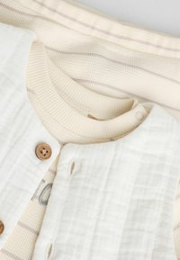 Conjunto para bebé de color claro que presenta un suave chaleco acolchado blanco con un botón de madera, superpuesto a una camiseta con rayas en crema y marrón.