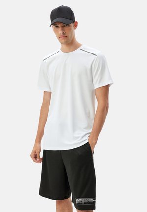 White short-sleeve athletic shirt with gray shoulder accents, paired with black shorts featuring a white text detail on the hem. Black cap.
