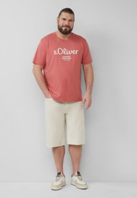 Rosa T-Shirt mit weißem Drucktext, kombiniert mit hellbeigen Shorts und weißen Sneakern. Einfaches, lässiges Design mit kurzen Ärmeln.