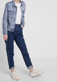 Jeansowa kurtka z dwoma kieszeniami na piersiach, jasno-niebieski kolor i kontrastowe paski na rękawach, zestawiona z ciemnymi spodniami o wysokim stanie i białymi sneakersami.