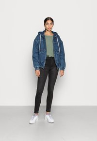 Chaqueta de mezclilla con capucha, dos bolsillos frontales, puños elásticos; lleva puesta sobre una camiseta verde y unos vaqueros ajustados negros. Zapatillas blancas completan el conjunto.