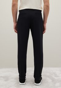 Pantaloni neri su misura con un fit slim, texture liscia, due tasche posteriori e risvolti, abbinati a sneakers nere su uno sfondo chiaro.