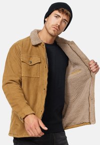 Bruine corduroy jas met een schapenvacht voering, een knoopjes borstzak en een zwarte gebreide beanie, open gedragen om de interieurtextuur en details te tonen.