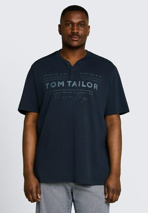 Man die een marineblauw Tom Tailor korte mouwen overhemd met knoopkraag en grijze broek draagt, staand tegen een effen lichte achtergrond.