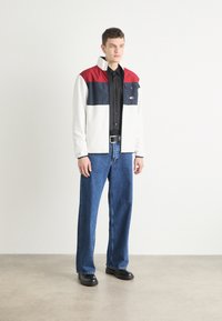 Giacca di pile bianca con blocchi di colore rosso e blu navy, camicia nera con bottoni, jeans a gamba larga blu e scarpe nere. Design semplice, vestibilità casual.