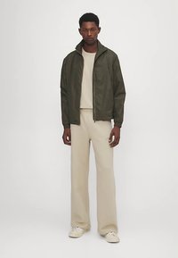 Oliven grøn let jakke med stående krave, lynlås foran, kombineret med en beige sweatshirt og bukser med afslappet pasform. Cremefarvede sneakers.