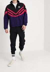 Adidas tepláková souprava s fialovou bundou, která má růžové diagonální pruhy, a černými kalhotami. Materiál vypadá lehký; součástí jsou černé a bílé sneakers.