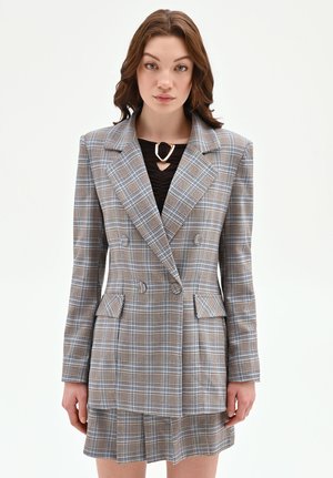 Femme portant un blazer croisé à carreaux beige et bleu avec une jupe assortie et un haut noir accompagné d'un collier triangulaire en or.