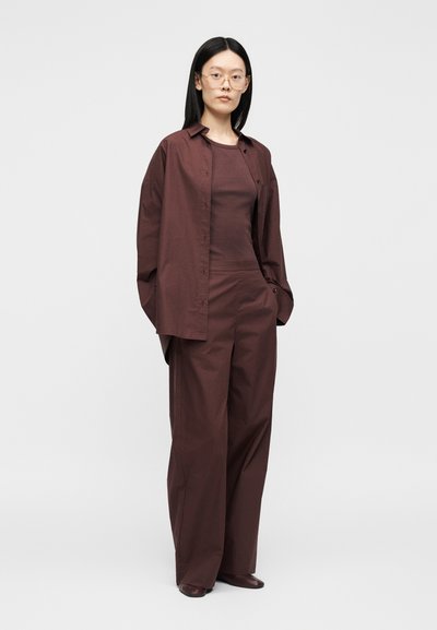 Femme aux cheveux noirs et raides, portant des lunettes, une chemise marron foncé ample, un pantalon large assorti et des chaussures foncées, debout les mains dans les poches.