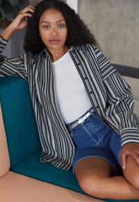 Chemise à rayures noir et blanc, associée à un débardeur blanc et un short en denim bleu, sur un canapé teal et rose.