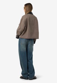 Vrouw in een oversized bruine jas met zwarte kraag, loszittende blauwe jeans, donkere schoenen en zonnebril, staand met de rug naar de camera op een witte achtergrond.