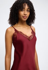 Γυναίκα με σγουρά μαλλιά που φοράει αμάνικο σκούρο κόκκινο τοπ με δαντελένια διακόσμηση, κοιτάζοντας την κάμερα σε απλό φωτεινό φόντο.