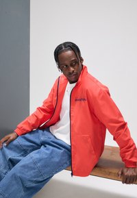 Röd zip-up jacka med marinblå detaljer och texten "Revolution"; lager på vit skjorta. Kombinerad med oversize blå denimbyxor.