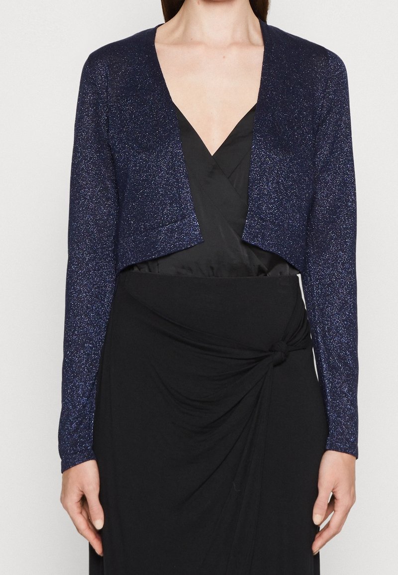 Cardigan pailleté bleu marine recadré avec manches longues, devant ouvert et tissu métallique texturé, associé à une robe portefeuille noire.