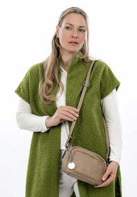 Borsa a tracolla in pelle tan con dettagli intrecciati e un ciondolo circolare. Indossata sopra una camicia bianca a maniche lunghe e un scialle verde texture.