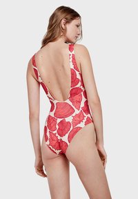 Un maillot de bain une pièce avec un dos en V profond, fabriqué en tissu léger avec une base blanche et des motifs de coquillage rouges.
