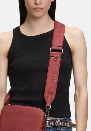Kobieta w czarnym topie bez rękawów, ozdobionym ćwiekami pasku i czerwoną torebką na ramię z szerokim paskiem z napisem "LIEBESKIND".